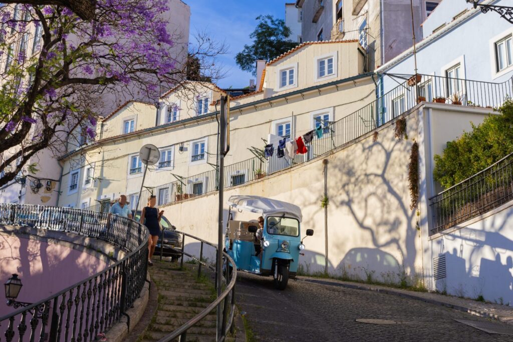 A light blue tuk-tuk descending a steep, narrow alley in Alfama, Lisbon