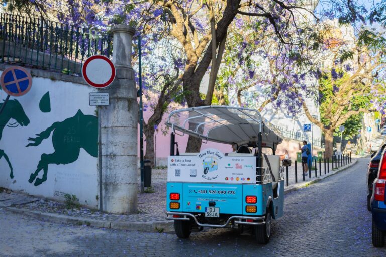 A Chico Chico Tours tuk-tuk parked beneath jacaranda trees beside a painted wall in Lisbon