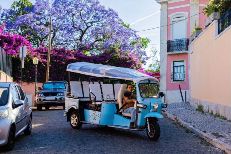 A light blue tuk-tuk on a Lisbon street beneath jacaranda blossom beside a pink building