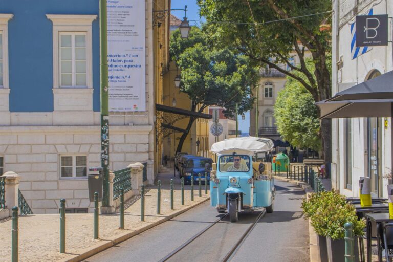 A light blue tuk-tuk crossing tram tracks on a quiet Lisbon street
