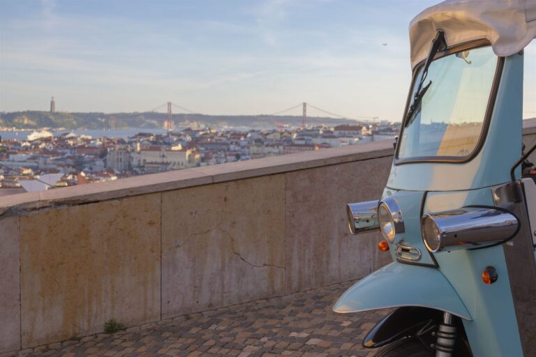 A light blue tuk-tuk parked at a Lisbon viewpoint wall above the city