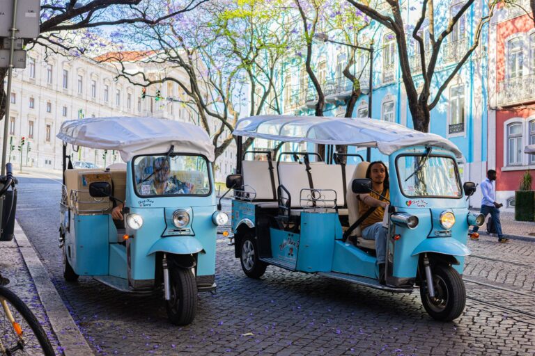 Two tuk-tuks carrying guests along a Lisbon avenue lined with jacaranda trees