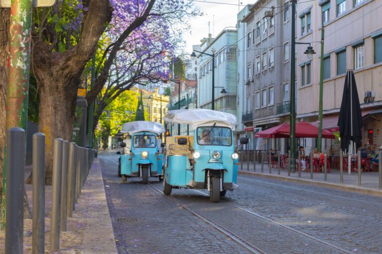 Two light blue Chico Chico Tours electric tuk-tuks driving down a Lisbon street lined with jacaranda trees in bloom